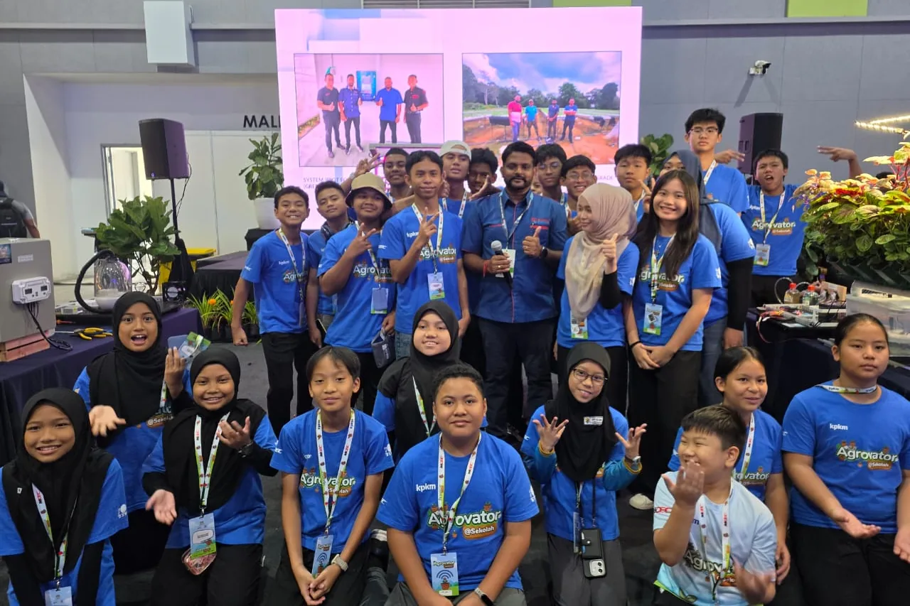 Students in Agrovator@Sekolah shirts gathered with a presenter holding a microphone, in front of a screen showing site photographs
